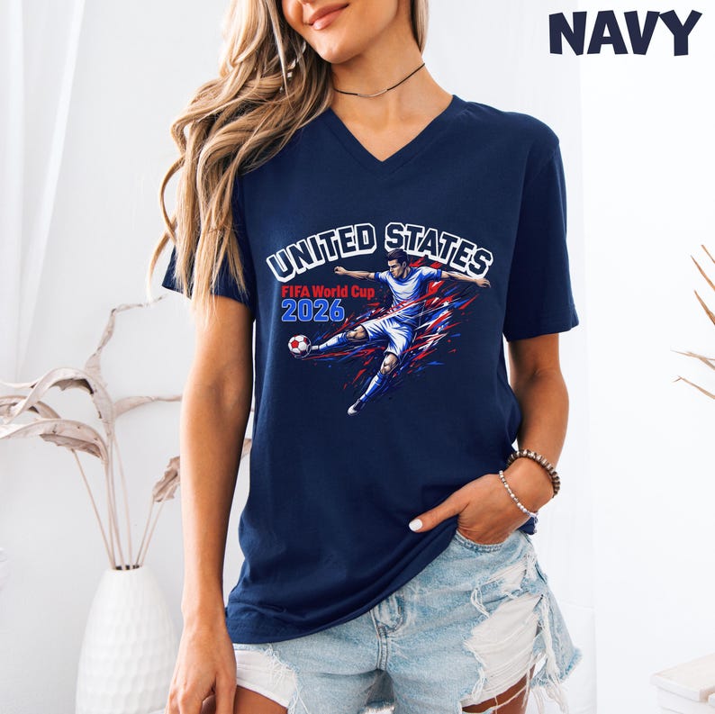 May include: Navy blue V-neck t-shirt featuring a soccer player graphic with the text "UNITED STATES FIFA World Cup 2026". The graphic has red, white, and blue accents. The word "NAVY" is printed at the top right.