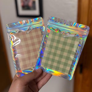May include: Two rectangular air fresheners with a gingham check pattern, one in light brown and one in sage green, each featuring a small white ribbon loop for hanging, individually sealed in clear iridescent holographic zipper pouches.