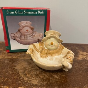 May include: A stone glaze snowman dish with a matching box. The dish is shaped like a snowman wearing a hat and scarf, holding a smaller snowman. The box reads "Stone Glaze Snowman Dish". The dish is beige and brown.