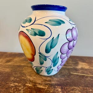 May include: A white ceramic vase with a blue rim, adorned with hand-painted fruit and foliage. The design includes a large orange fruit, purple grapes, and green leaves with blue vines. The vase sits on a wooden surface.