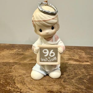 May include: A ceramic figurine of a child angel with a halo, holding a sign that reads "96 Peace on Earth." The angel wears a white robe, pink accents, and light blue pants. The figurine is on a wooden surface.