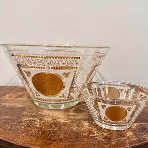 May include: Two clear glass bowls with gold detailing. The larger bowl has a wide rim and a decorative pattern with a central gold medallion. The smaller bowl mirrors the design. Both bowls sit on a wooden surface.