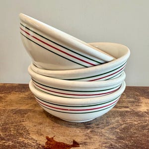 May include: A stack of four off-white ceramic bowls, each featuring a decorative pattern of two thin horizontal stripes in red and green encircling the exterior. The bowls have a smooth, rounded rim and a deep, curved basin for serving food.
