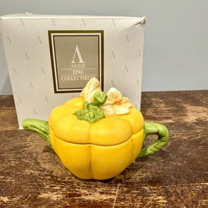 May include: A yellow ceramic teapot shaped like a pumpkin, with a green handle and spout. The lid is adorned with a floral design. The teapot is in front of a white box with the text "AVON FINE COLLECTIBLES".