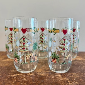 May include: Set of six clear drinking glasses. Each glass features a birdhouse design with red hearts, green foliage, and a small sign that reads "Keep". The glasses are arranged on a wooden surface.
