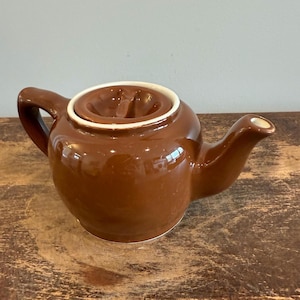 May include: A brown ceramic teapot with a rounded body, a curved spout, and a handle. The lid has a recessed area with a white rim. The teapot is sitting on a wooden surface.