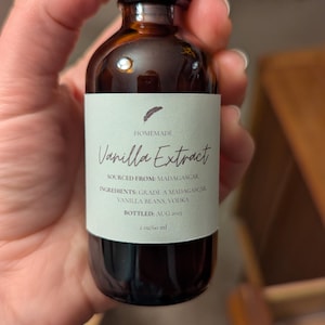 May include: A small brown glass bottle of homemade vanilla extract. The label reads "Vanilla Extract" and lists the ingredients as Madagascar vanilla beans and vodka. The bottle is 2 oz / 60 ml and bottled in August 2025.
