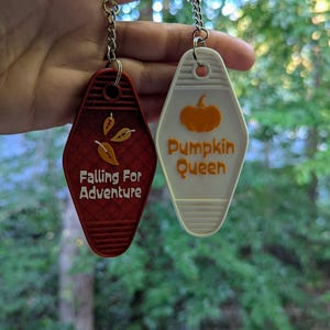 May include: Two motel-style keychains. One is red with the text "Falling For Adventure" and gold leaves. The other is white with an orange pumpkin graphic and the text "Pumpkin Queen". Both have silver keychains.