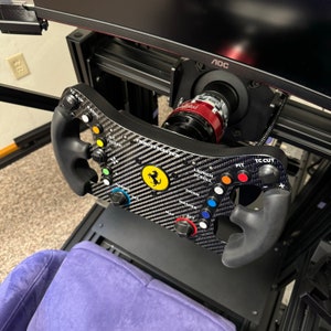 Puede incluir: Un volante de carreras de fibra de carbono con el logotipo de Ferrari y m&uacute;ltiples botones de colores. El volante est&aacute; montado en un marco negro con un monitor en el fondo. El volante tiene empu&ntilde;aduras negras y est&aacute; dise&ntilde;ado para simulaci&oacute;n de carreras.