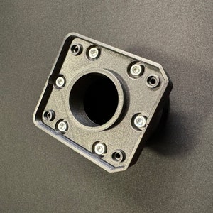 Puede incluir: Un componente negro de forma geom&eacute;trica con una abertura circular central. Cuenta con m&uacute;ltiples orificios para tornillos alrededor del per&iacute;metro, cada uno con un tornillo plateado. El objeto est&aacute; hecho de un material texturizado y descansa sobre una superficie oscura.