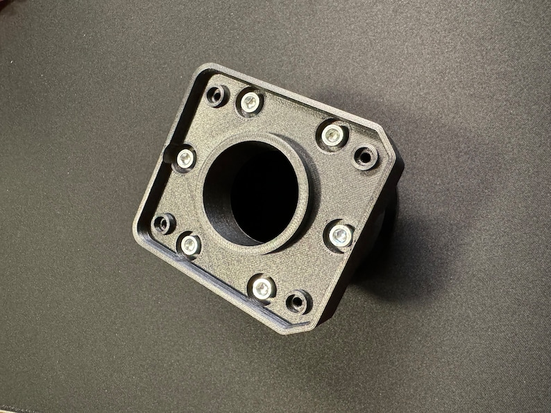 Puede incluir: Un componente negro de forma geom&eacute;trica con una abertura circular central. Cuenta con m&uacute;ltiples orificios para tornillos alrededor del per&iacute;metro, cada uno con un tornillo plateado. El objeto est&aacute; hecho de un material texturizado y descansa sobre una superficie oscura.