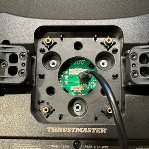 Puede incluir: Primer plano de una base de volante de carreras negra Thrustmaster F488-GT3-WW. La base tiene una placa de circuito verde central con un cable negro. La base tiene varios puntos de montaje y el logotipo de Thrustmaster.