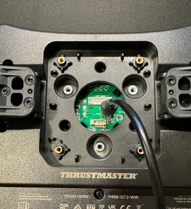 Puede incluir: Primer plano de una base de volante de carreras negra Thrustmaster F488-GT3-WW. La base tiene una placa de circuito verde central con un cable negro. La base tiene varios puntos de montaje y el logotipo de Thrustmaster.