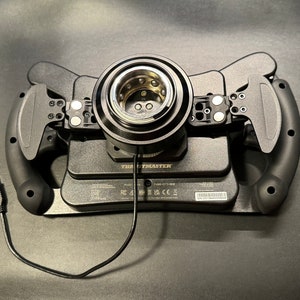 Puede incluir: Volante de carreras negro con levas y cable USB. El volante tiene un cubo central con un anillo de metal plateado y est&aacute; montado sobre una base negra. La marca "Thrustmaster" es visible en la base.