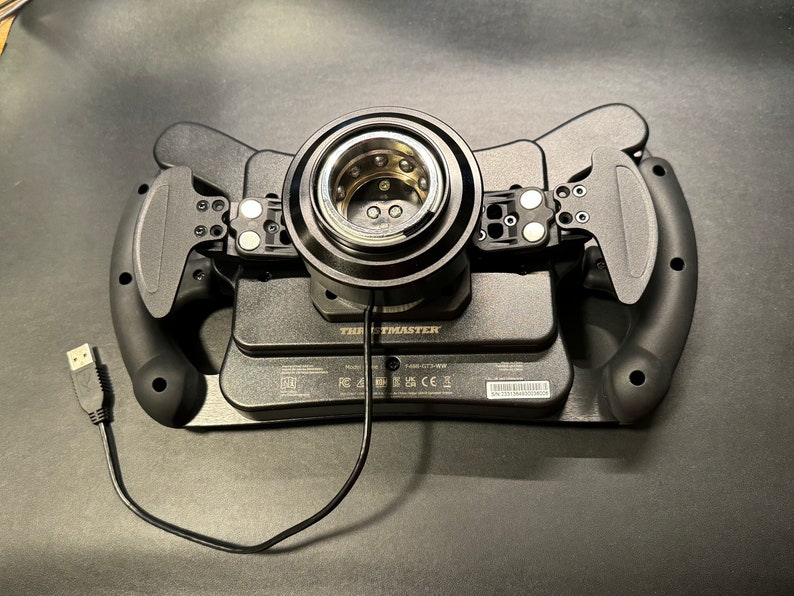 Puede incluir: Volante de carreras negro con levas y cable USB. El volante tiene un cubo central con un anillo de metal plateado y est&aacute; montado sobre una base negra. La marca "Thrustmaster" es visible en la base.