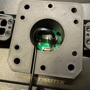 Puede incluir: Primer plano de un componente gris de forma cuadrada con una abertura circular central. Un cable negro se extiende desde la abertura, conectado a una placa de circuito verde. El componente est&aacute; montado sobre una superficie negra, con la palabra "THRUSTMASTER" visible.