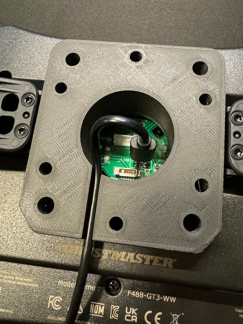 Puede incluir: Primer plano de un componente gris de forma cuadrada con una abertura circular central. Un cable negro se extiende desde la abertura, conectado a una placa de circuito verde. El componente est&aacute; montado sobre una superficie negra, con la palabra "THRUSTMASTER" visible.