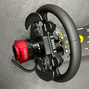 Puede incluir: Un volante de carreras negro con un cubo central rojo, con el texto "MOMO ITALY". El volante tiene levas de cambio y m&uacute;ltiples botones de colores. El fondo es una superficie gris oscuro.