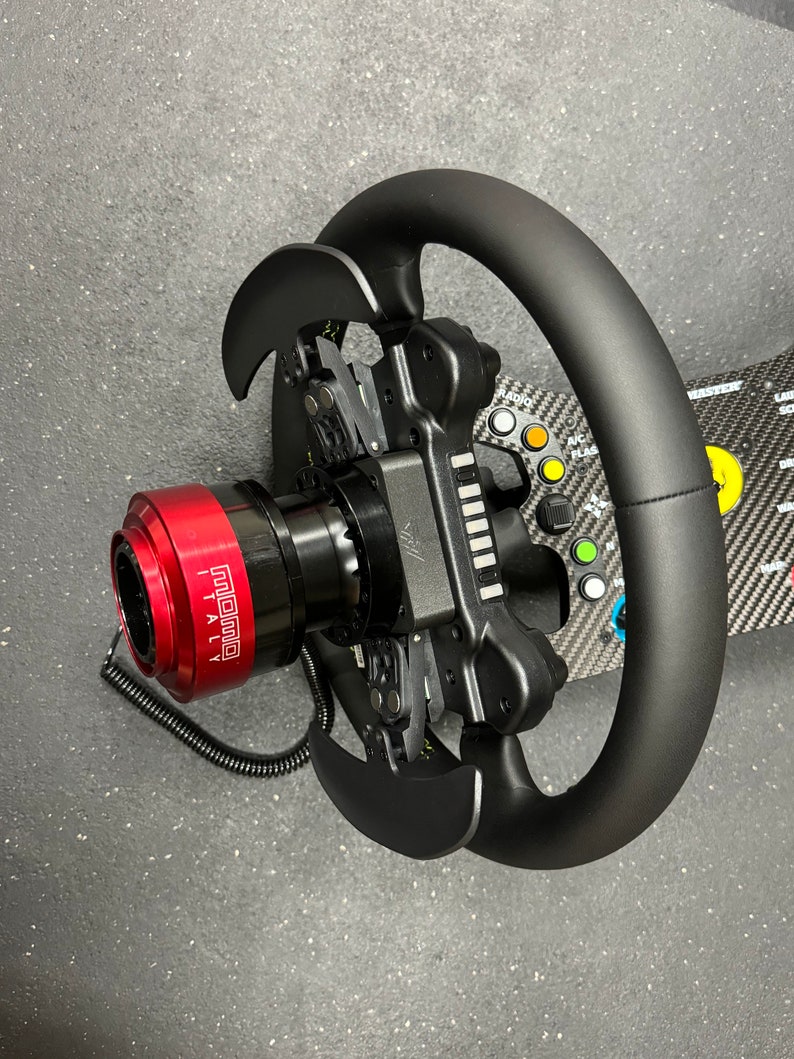 Puede incluir: Un volante de carreras negro con un cubo central rojo, con el texto "MOMO ITALY". El volante tiene levas de cambio y m&uacute;ltiples botones de colores. El fondo es una superficie gris oscuro.
