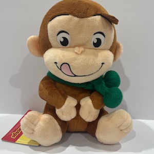 May include: A plush monkey with brown fur, a tan face, and a green scarf. The monkey has a smiling expression with its tongue sticking out. The toy is sitting down and has a tag attached.