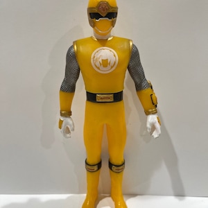 May include: Yellow action figure of a Power Ranger with a gold helmet, a white face mask, and a gold belt. The figure has a white symbol on the chest and is wearing silver armor on the arms.
