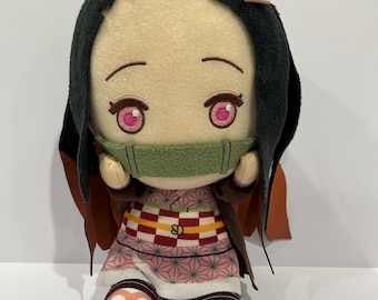 Op de afbeelding: Een pluche pop van een personage met zwart haar, roze ogen en een groene mondbeschermer. De pop draagt een roze en witte kimono met een bruine jas en bruine en bruin-beige schoenen. Een roze haaraccessoire zit op het hoofd van de pop.