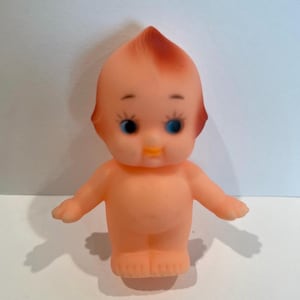 May include: A small, pink rubber doll with a red and orange mohawk, blue eyes, and a yellow smile.