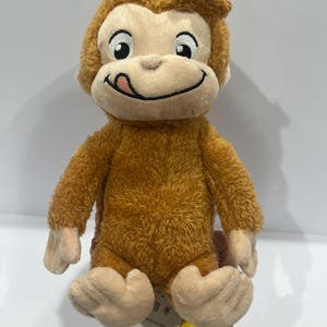 May include: A soft toy monkey with brown fur and a light tan face, featuring a smiling expression with its tongue out. The plush monkey has a cuddly appearance. A yellow tag attached reads "Made in China".