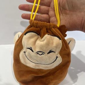 May include: A brown plush drawstring bag shaped like a smiling monkey face. The bag has a light tan face, brown ears, and a yellow drawstring. The bag is held up by a hand.