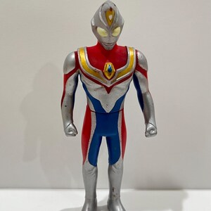 May include: A silver, red, and blue action figure of a superhero with a diamond-shaped chest emblem. The figure has a pointed head with a silver crest and yellow eyes.