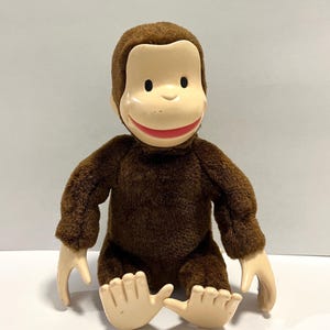 May include: A plush toy of a monkey with brown fur and a smooth, light-colored plastic face. The monkey has black eyes, a wide red-painted mouth, and light-colored plastic hands and feet. The figure is seated in an upright position.
