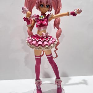 Op de afbeelding: Een roze anime-stijl figuur met lange, krullende paardenstaarten en blauwe ogen. Het figuur draagt een gelaagde roze en witte jurk, roze laarzen en armbanden. Het houdt een roze accessoire in één hand en heeft de andere arm uitgestrekt.