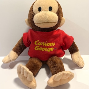 May include: A plush monkey toy with brown fur and a tan face, ears, hands, and feet. The monkey is wearing a red short-sleeved t-shirt with the text Curious George in yellow lettering on the front. The toy has black plastic eyes.