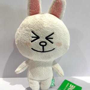 May include: A white plush rabbit toy with pink inner ears, rosy cheeks, and closed eyes. The toy has a black mouth and whiskers. A green tag with the word "LINE" is attached. The toy is a LINE Friends character.