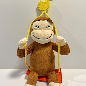 May include: A brown plush monkey with a cream-colored face, hands, and feet, sitting on a small orange swing. The swing is suspended by yellow ropes attached to a yellow plastic banana-shaped hook at the top for hanging display.