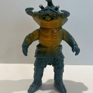 May include: A vintage action figure of a creature with a crab-like head, yellow torso, and blue-green limbs. The figure has multiple eyes and a textured surface, standing upright. This collectible toy is likely from a science fiction or monster-themed series.