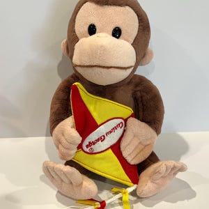 May include: A plush Curious George monkey doll, brown with a tan face and hands, holding a yellow and red kite. The kite has the words "Curious George" printed on it. The doll is sitting.