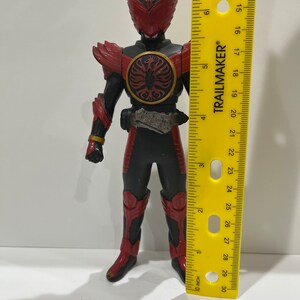 May include: A red and black action figure with a red helmet and wings. The figure is holding a sword and has a belt with a buckle. The figure is standing on a yellow ruler that is marked in inches.