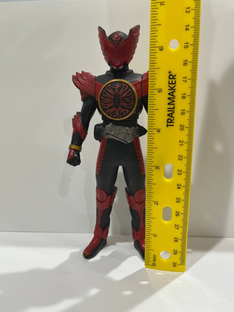 May include: A red and black action figure with a red helmet and wings. The figure is holding a sword and has a belt with a buckle. The figure is standing on a yellow ruler that is marked in inches.