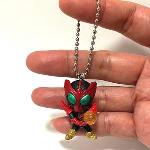 Op de afbeelding: Een kleine, gestileerde figuur-sleutelhanger met een personage met een zwart-rode helm, grote groene ogen en gouden laarzen. Het figuurtje is bevestigd aan een zilverkleurige metalen kralenketting en houdt een doorschijnend oranje bolvormig object vast.