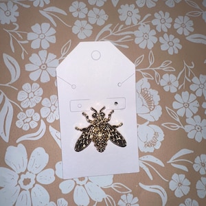 May include: A gold-colored bee pin on a white tag. The bee pin has detailed wings and body. The background is a peach color with a white floral pattern.