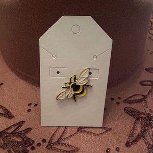 May include: A bee-shaped enamel pin with black, yellow, and white details, displayed on a white card. The pin features a detailed design of a bee with outstretched wings. The background is a textured, brown surface.