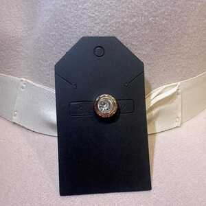 May include: A gold-toned button with a clear, round gem is attached to a black tag. The tag is placed on a light pink hat with a cream-colored band. The button has a decorative, textured border.