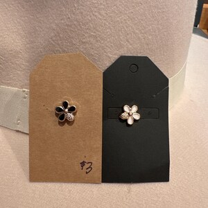 May include: Two flower-shaped stud earrings, one black and gold, the other white and gold, displayed on small tags. The earrings are set against a neutral-toned hat, showcasing the jewelry's design and color contrast.