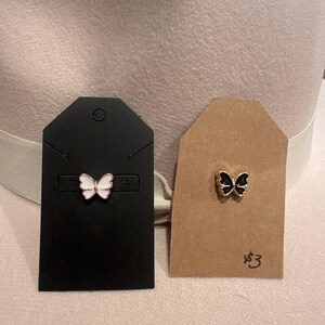 May include: Two butterfly-shaped pins on display cards. One pin is white and pink, the other is black. The cards are black and tan, and are displayed against a light-colored hat.