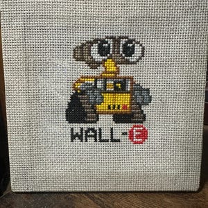 May include: A cross-stitch of the WALL-E character from the animated film. The robot is depicted in yellow, black, and brown, with the name "WALL-E" stitched below in black and red. The background is a light beige fabric.