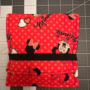 May include: Red fabric pouch with a black elastic band. The pouch has a polka dot pattern and features Minnie Mouse characters with the word "Minnie" printed on it.