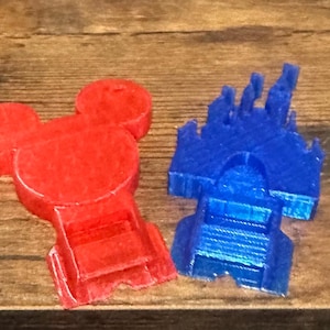 May include: Two 3D-printed plastic shapes: a red Mickey Mouse head and a blue castle. The Mickey Mouse shape has a rounded head with two large ears. The castle has a detailed facade with turrets and a central archway.