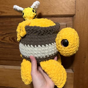 May include: A handmade, plush bee-themed toy. The toy features a yellow body, brown and white striped pot, and a bee with white and black accents. The bee is holding a pot of yellow honey. The toy is made of soft, textured yarn.