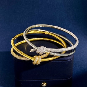 May include: Three gold and silver bangle bracelets with a knot design. The bracelets are encrusted with small, clear stones.